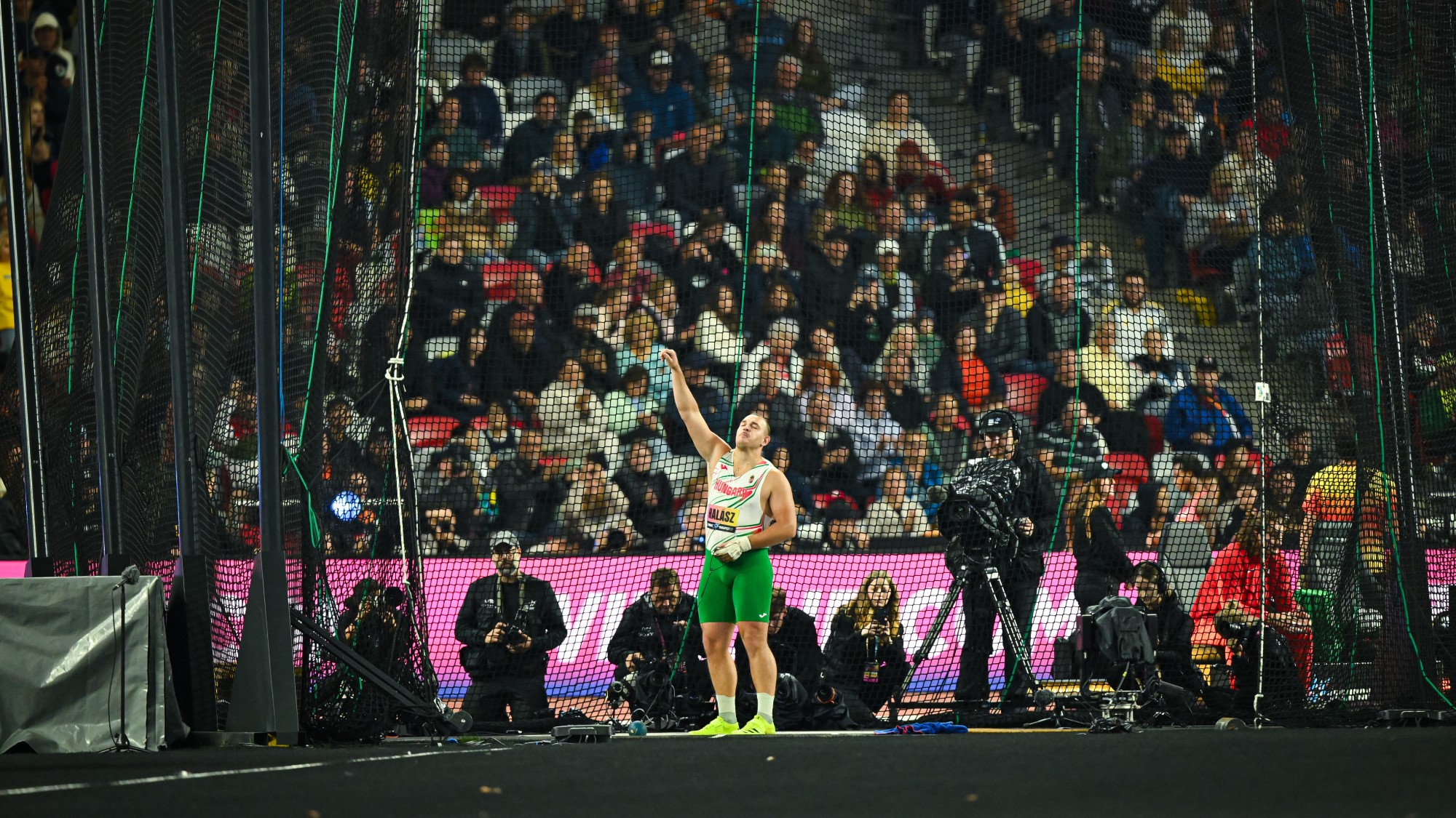 Hammer thrower in green shorts and white jersey inside a netted throwing cage, raising his right arm as photographers and a cheering crowd watch in the stands.
