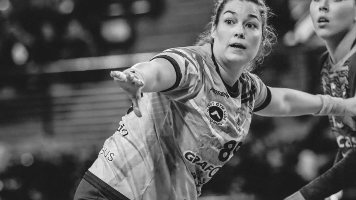 Female athlete in a jersey reaching forward with outstretched arm during an indoor team sport moment, likely handball or volleyball, in a gym setting